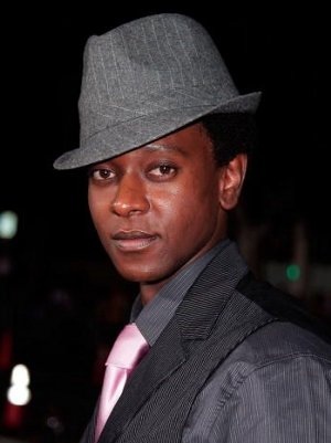 A torso shot of Edi Gathegi - a black man with short, very dark brown hair. He is standing side on and turning to face the camera. He is wearing a grey suit and hat, and a pale pink tie. 